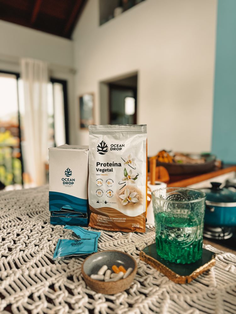 Suplementos Ocean Drop dispostos sobre uma mesa, com destaque para a proteína vegetal sabor baunilha e cápsulas ao lado de um copo verde.