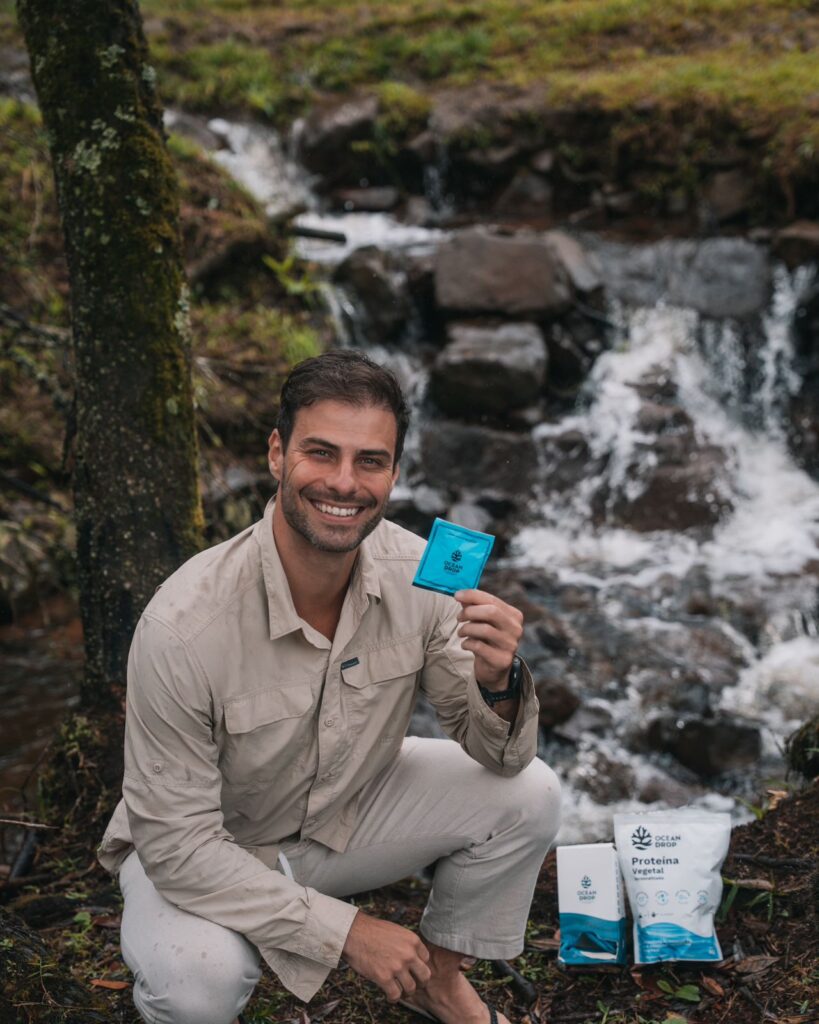 Lucas Malvacini segura um sachê da Ocean Drop em ambiente natural, com produtos da marca ao lado e uma cachoeira ao fundo.