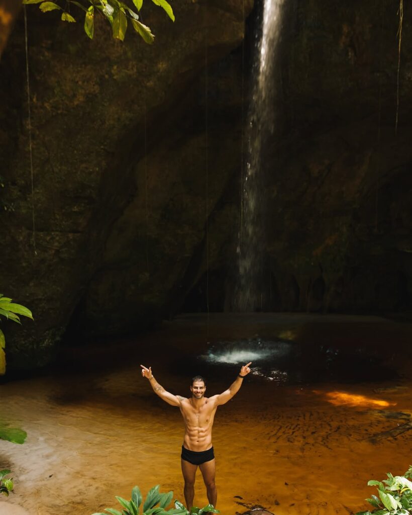 Homem em boa forma física posando sorridente sob uma cachoeira em meio à natureza, com os braços erguidos.