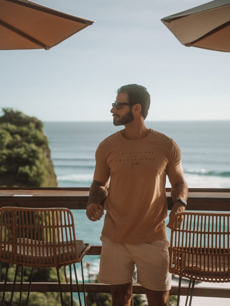 Homem de camiseta bege e óculos escuros em um deck com vista para o mar, em um ambiente iluminado e natural.