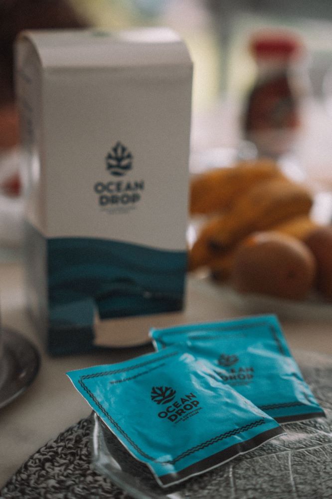 Embalagens da Ocean Drop sobre uma mesa de café da manhã, com sachês azuis e frutas ao fundo.