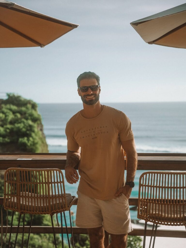 Homem de camiseta bege e óculos escuros em um deck de frente para o mar, sob luz natural.
