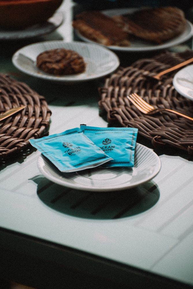 Sachês azuis da Ocean Drop sobre um prato, dispostos em uma mesa de café da manhã com louças e alimentos.